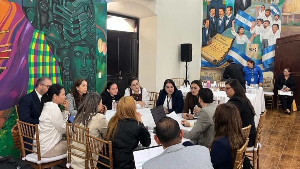 Professionals seated around a round table in a room with a vibrant mural, reviewing documents and laptops.