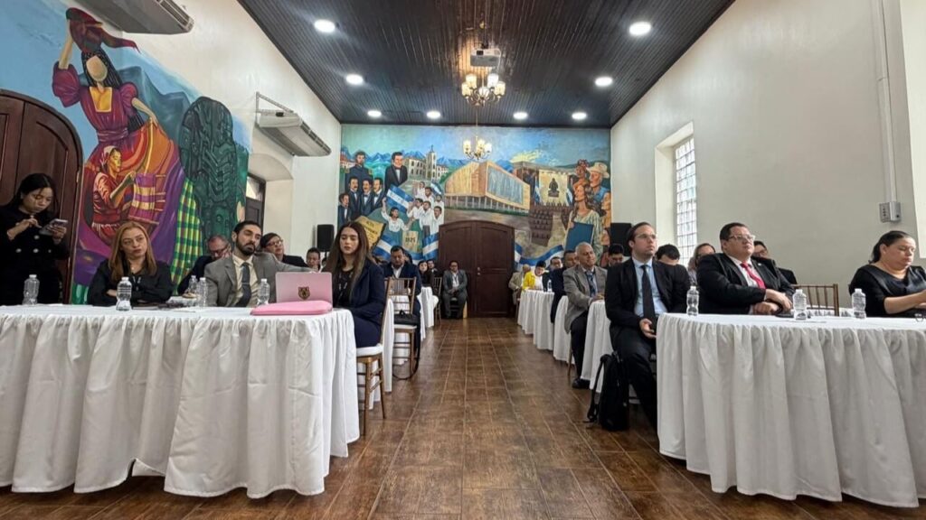 Formal meeting or conference with attendees seated at long white-clothed tables in a mural-filled room, with a pink laptop on the front table and bright ceiling lights.