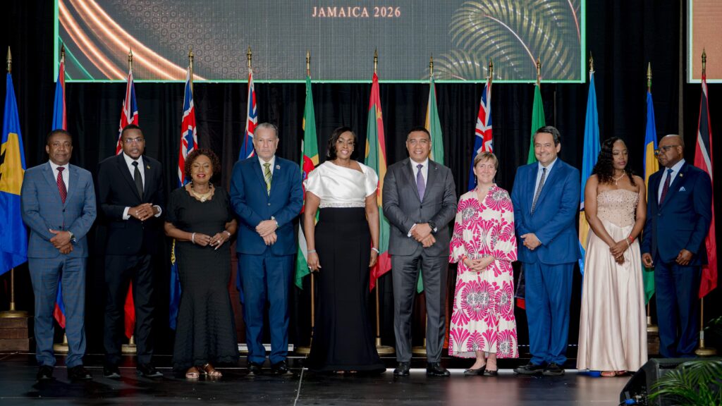 Group of diverse officials in formal attire standing on a stage with flags behind them, Jamaica 2026 banner above.