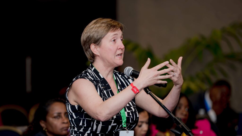 Woman on stage speaking into a microphone, gesturing with her hands while addressing an audience.