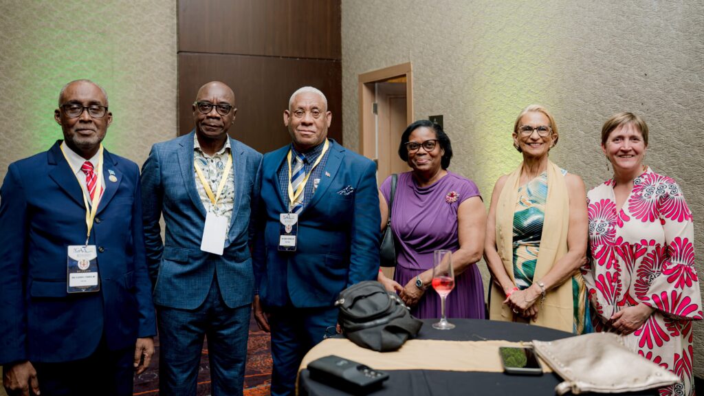 Six adults in colorful formal attire pose together at a reception, wearing event badges, with a table and wine glasses in the foreground.