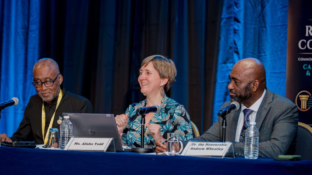 Panel discussion with three speakers at a conference table; center woman smiles, flanked by two men, microphones and nameplates visible (Ms. Alisha Todd).