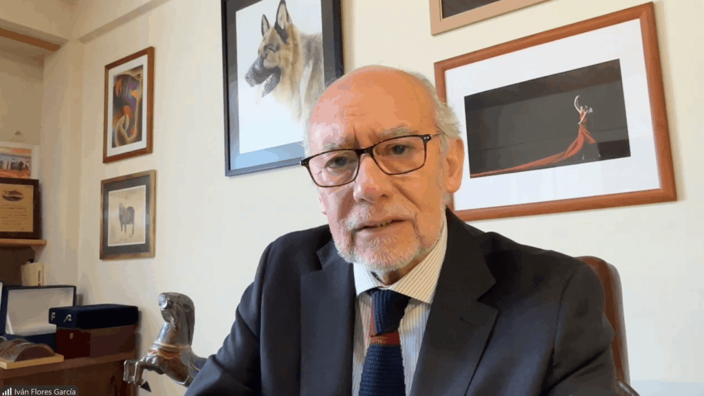 Older man wearing glasses and a dark suit speaks during a video call in a home office with framed art on the wall behind him.