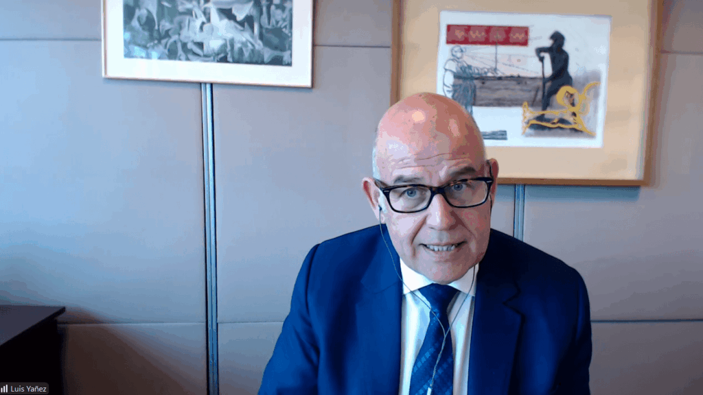 Bald man wearing glasses in a blue suit speaks during a video conference in a modern office setting.