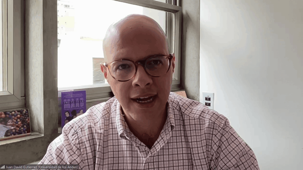 Man with glasses in a checkered shirt speaks during a video call in a bright office inside a windowed room.