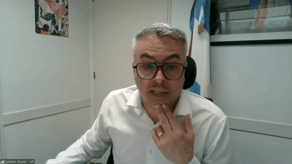 Man with short gray hair and glasses, wearing a white shirt, speaks during a video call from an office setting with a flag in the background.