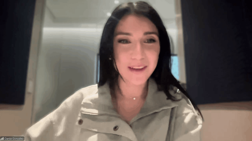 Woman with shoulder-length dark hair speaks to camera in a well-lit room during a video call, wearing a light-colored jacket.