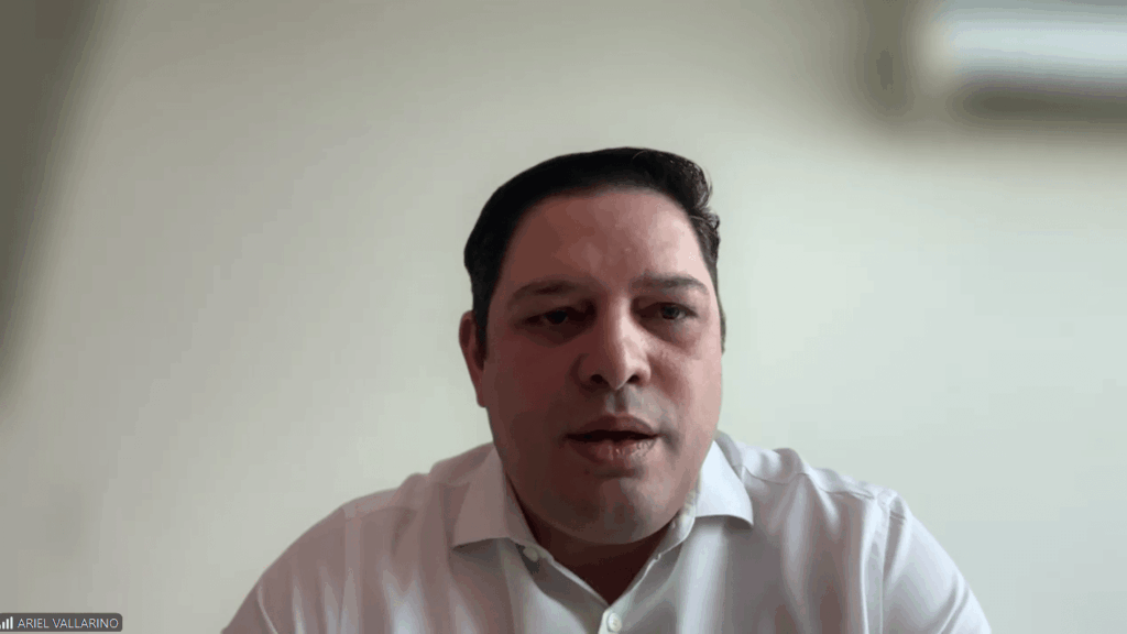 Man in a white shirt speaks during a video conference, facing the camera with a neutral background.