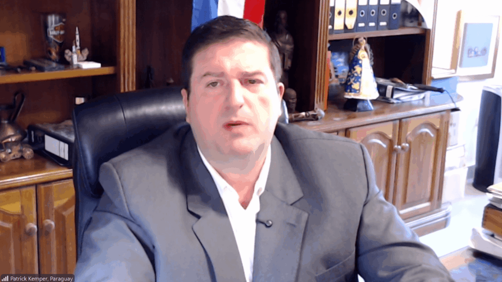 Man in a gray suit speaking during a video call from a formal office with wooden shelving and a flag in the background.