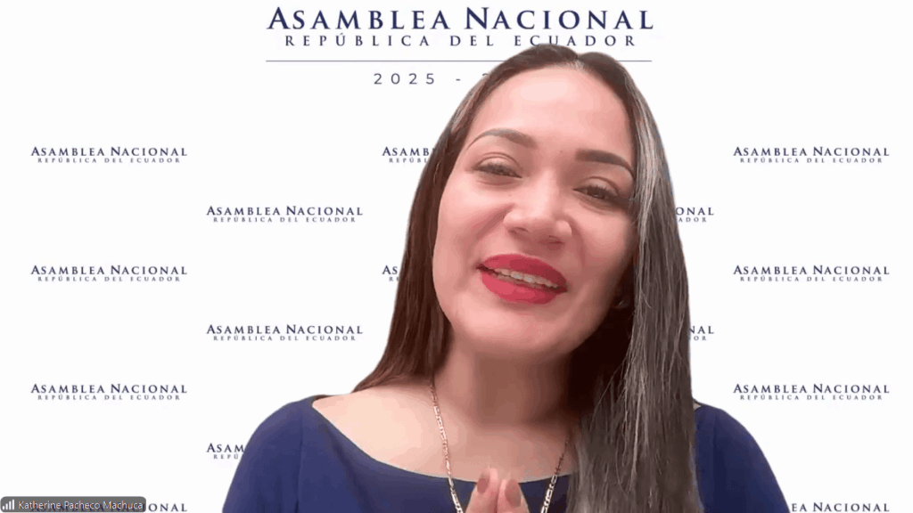 Woman with long dark hair in a blue top smiles at the camera during a video call, standing in front of an 'Asamblea Nacional—República del Ecuador' backdrop.