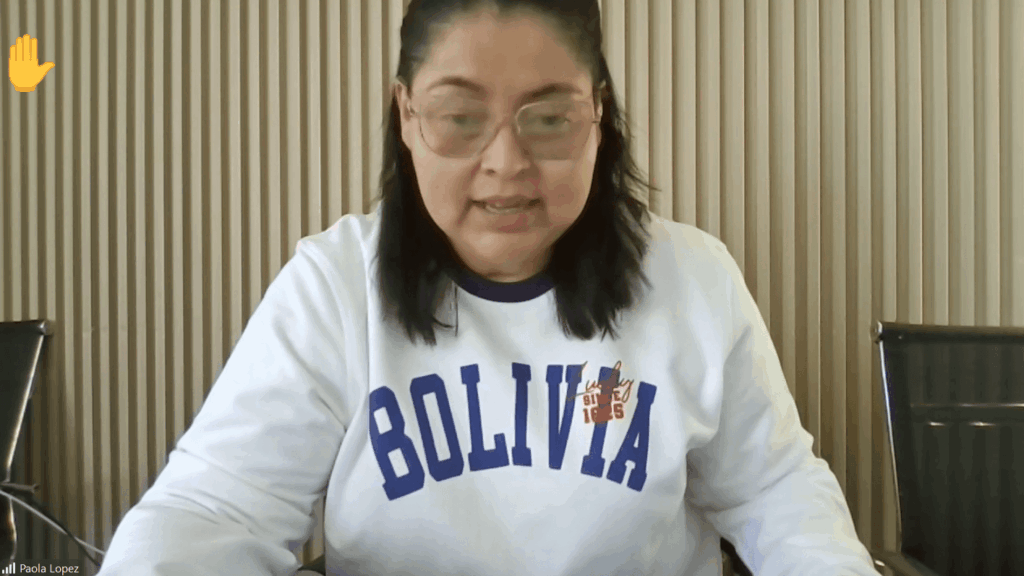 Woman with shoulder-length dark hair and glasses sits at a desk in a video call, wearing a white sweatshirt that reads 'BOLIVIA'.