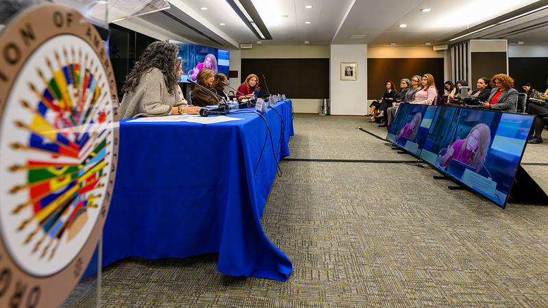 Panel of speakers seated at a long blue-draped table in a conference room; screens show remote participants.