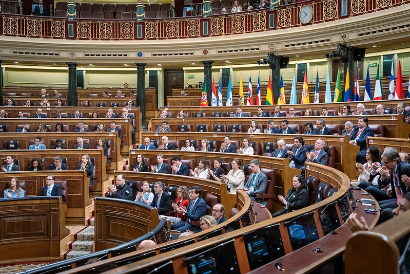 Parliament chamber with lawmakers seated in curved wooden benches, flags along the wall.