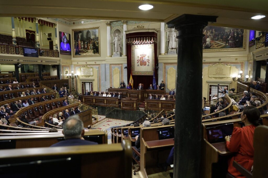 Wide view of an ornate parliamentary chamber with lawmakers seated in curved benches around a central dais, flags and a crest behind the speakers.