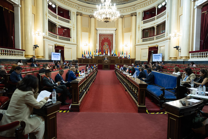 Grand parliamentary chamber with a central podium, red carpet, and delegates seated at long desks facing the speaker's dais.