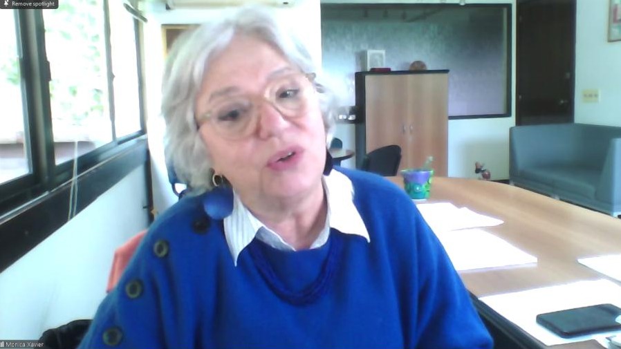 Older woman with white hair and glasses speaks during a video call in a bright office, sitting at a table with papers in front of her.