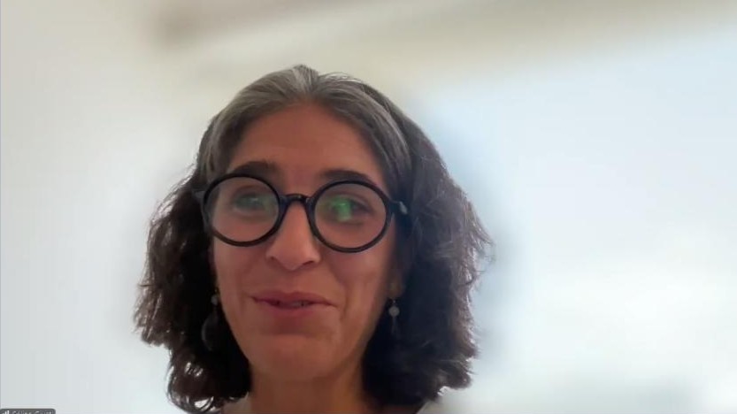Woman with shoulder-length dark hair wearing large round glasses and earrings, smiling on a video call. (informational)