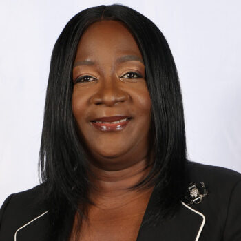 Portrait of a smiling Black woman with straight black hair, wearing a black blazer with white piping and a decorative brooch at the shoulder.
